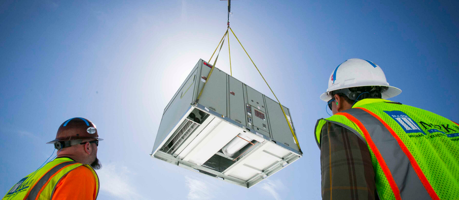 Two construction workers watch as a large HVAC unit is lowered by a crane under a bright sky.