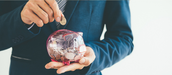Man in blue suit holds coin above clear piggy bank that is already half way full of money.