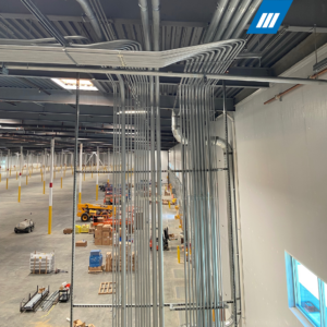 Industrial warehouse interior with an extensive network of metal conduit pipes along the ceiling and walls, forklifts, pallets, boxes, and yellow support columns.