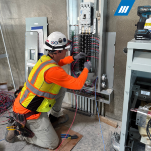 Brent Davidson wearing a safety helmet, glasses, gloves, and a high-visibility vest, kneeling and working on an electrical panel with tools and wires.
