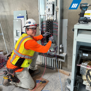 Brent Davidson wearing a safety helmet, glasses, gloves, and a high-visibility vest, kneeling and working on an electrical panel with tools and wires.